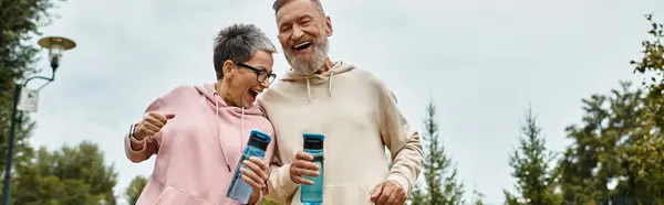 A mature couple enjoys a playful walk together in the park, smiling and holding water bottles close. — Stock Photo