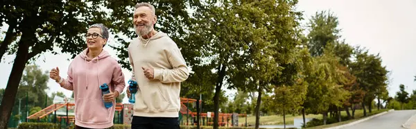 A loving couple exercises together, smiling as they jog through a beautiful, green park. — Stock Photo