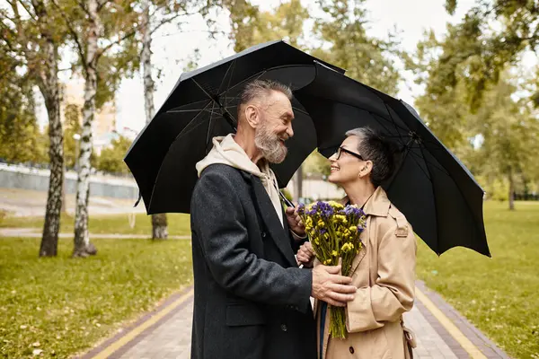 A loving couple enjoys a romantic moment under an umbrella while holding flowers in a park. — Stock Photo