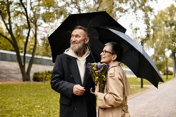 A couple strolls in a green park, sharing joy under umbrellas with flowers in hand. — Fotografia de Stock