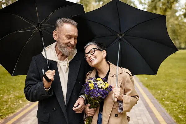 Ein Paar genießt einen fröhlichen Spaziergang im Park, lächelt, während es Sonnenschirme und Blumen in der Hand hält. — Stockfoto