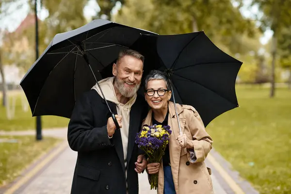 A mature couple enjoys a joyful stroll in the park, embracing each other with flowers in hand. — Stock Photo