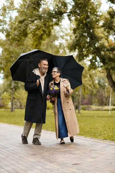 A mature couple strolls happily through a lush park while sharing umbrellas in light rain. — Stock Photo