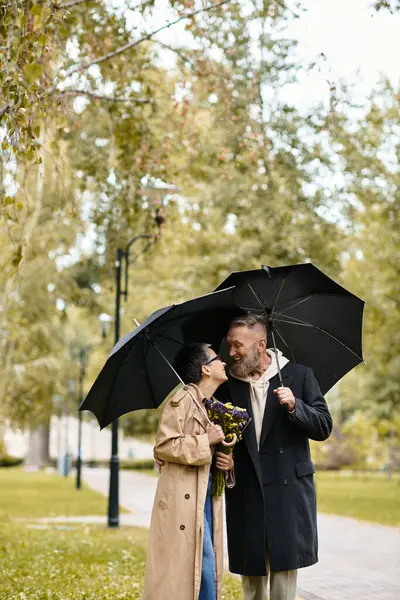 A mature couple enjoys laughs and kisses under a black umbrella in a serene park. — Stock Photo