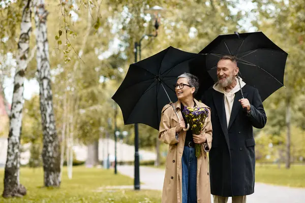 A loving couple strolls through a park, sharing umbrellas while holding vibrant flowers and smiling. — Stock Photo