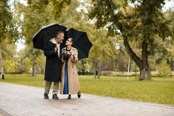 A loving senior couple strolls through a lush park, sharing umbrellas and joy in the rain. — Stock Photo