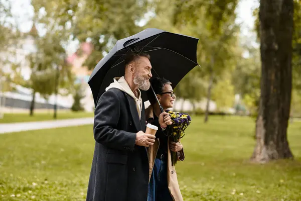 A loving couple enjoys a rainy walk in the park, smiling as they share a cozy umbrella together. — Stock Photo