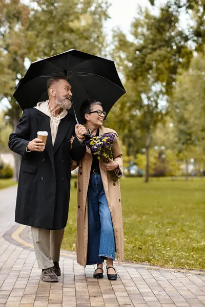 A loving couple strolls together in a vibrant park, enjoying coffee while sharing a cozy umbrella. — Stock Photo