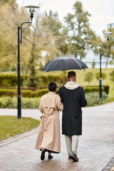 A loving couple walks hand in hand beneath an umbrella, sharing joyful moments in a tranquil park. — Stock Photo