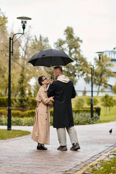 A loving couple embraces under an umbrella, sharing warmth and joy in the park during light rain. — Stock Photo