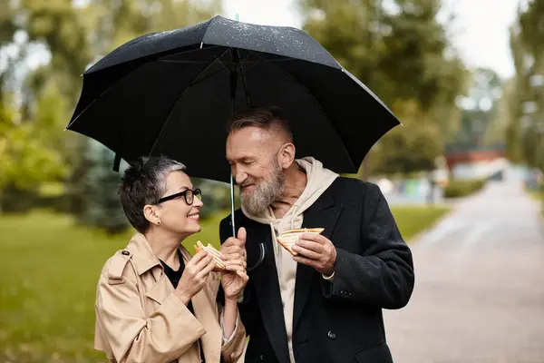 A loving couple enjoys sandwiches together while staying dry under a shared umbrella during a rain. — Stock Photo