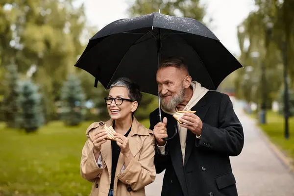 A loving couple enjoys snacks while walking together on a rainy day, sharing smiles and affection. — Stock Photo