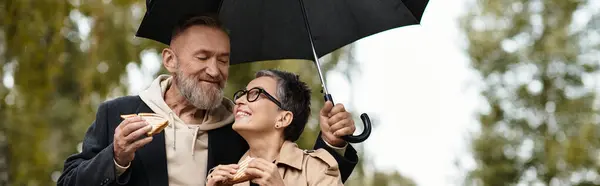 A loving couple stands close together, sharing sandwiches and smiles under an umbrella outdoors. — Stock Photo