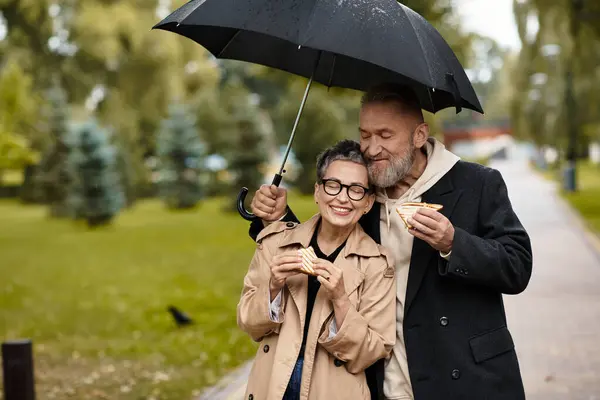 A loving couple enjoys a rainy day in the park while sharing delicious sandwiches and laughter. — Stock Photo