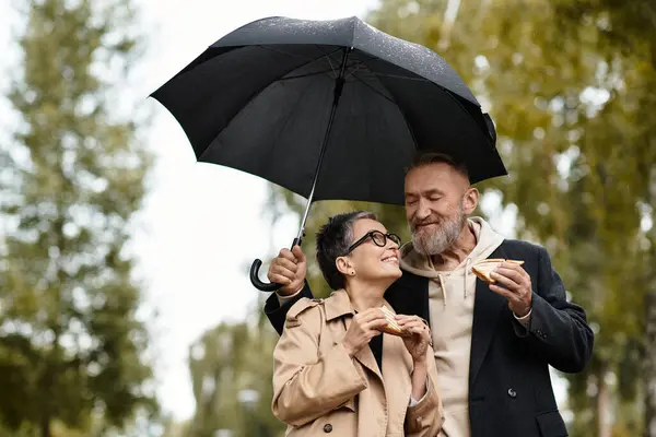 A loving couple enjoys sandwiches under an umbrella while surrounded by trees on a rainy day. — Stock Photo