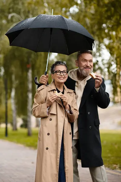 A loving couple enjoys snacks while standing under an umbrella on a rainy day in the park. — Stock Photo