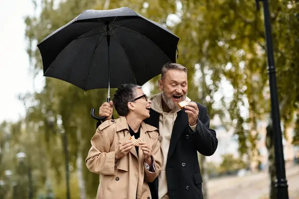 A mature couple enjoys a delightful snack, laughing together while holding an umbrella in the park. — Stock Photo