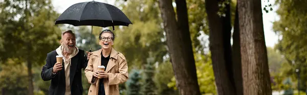 A loving couple strolls through a park, sipping coffee and laughing under an umbrella. — Stock Photo