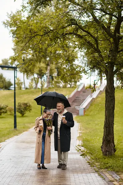 Mature couple enjoys a romantic walk under an umbrella, sharing smiles and love in a lush park. — Stock Photo
