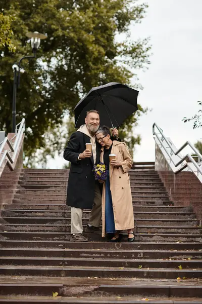 A loving couple walks down the steps on a rainy day while sharing an umbrella and warm drinks. — Stock Photo