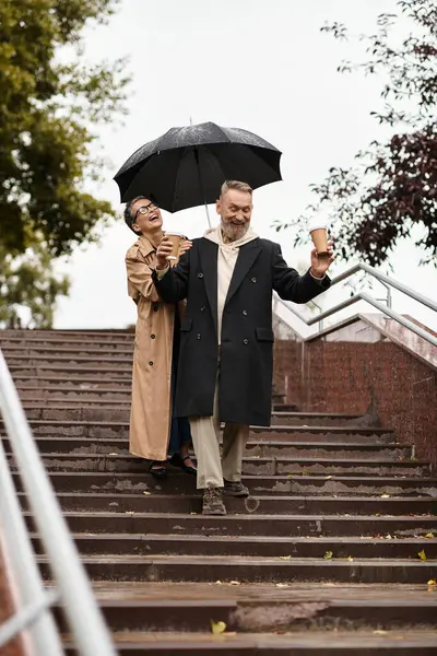 A couple laughs under a black umbrella, walking hand in hand down the rainy stairs. — Stock Photo