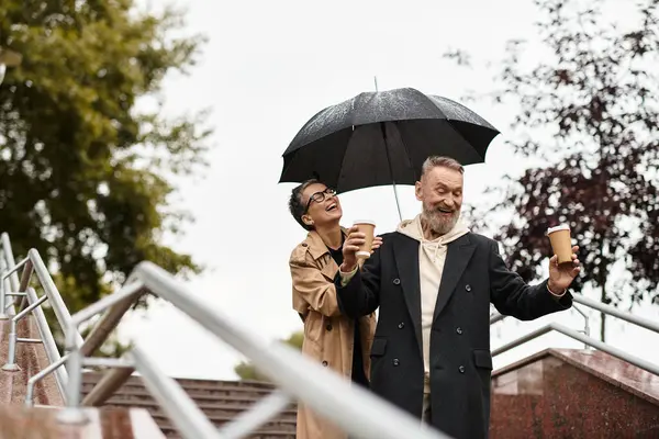 A couple laughs under an umbrella, sipping coffee while walking in the rain. — Stock Photo