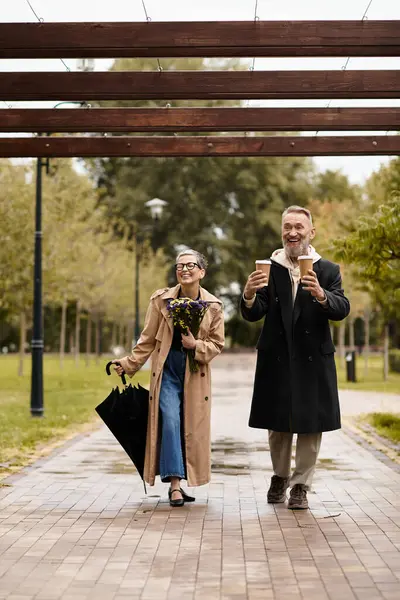 A joyful couple walks hand in hand in a lovely park, enjoying warm drinks and each others company. — Stock Photo