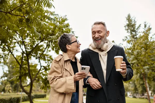 En un parque tranquilo, una pareja cariñosa comparte la risa mientras camina de la mano y disfruta del café. - foto de stock