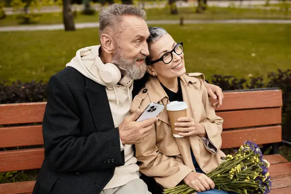 A mature couple relaxes on a park bench, sharing laughter, coffee, and a bouquet of flowers. — Stock Photo
