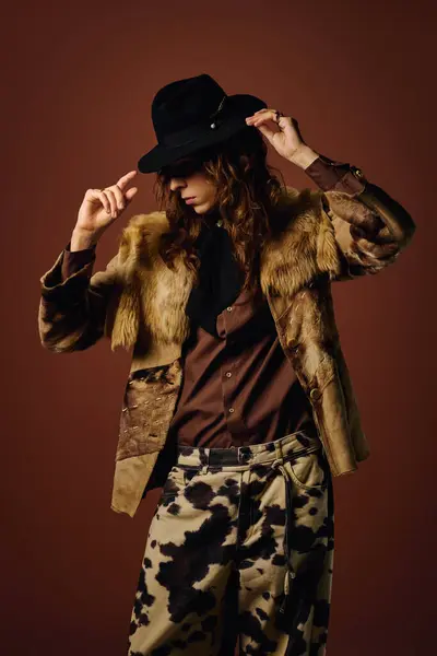 A handsome young man adjusts his black hat while wearing a fashionable eclectic outfit in a studio. — Stock Photo