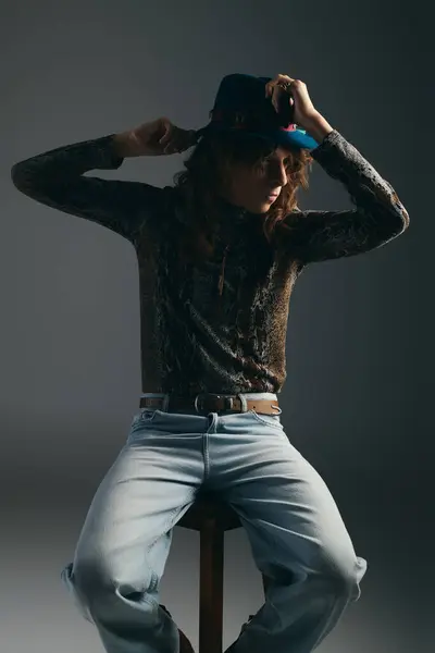 A young, handsome man poses elegantly on a stool, adjusting his stylish hat with charisma. — Stock Photo