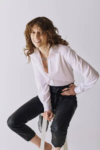Young man wearing a white shirt and black pants smiles while sitting on a stool in a studio. — Stock Photo