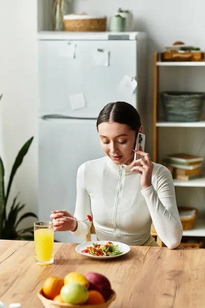 Una mujer joven disfruta de una colorida comida saludable en su mesa de la cocina mientras está en una llamada telefónica. - foto de stock