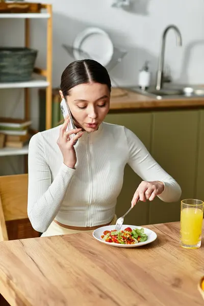 A young woman savors a nutritious meal in her cozy kitchen, talking on her phone. — Stock Photo