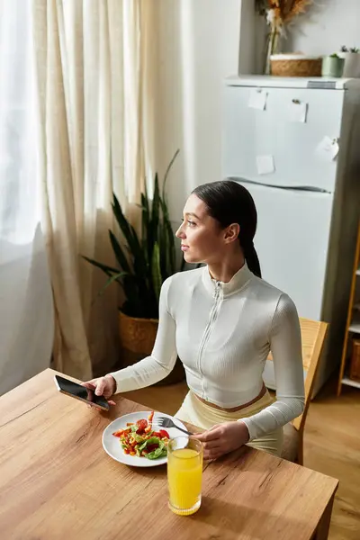 A young woman has a healthy breakfast of salad and a drink at her kitchen table. — Stock Photo
