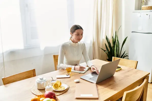 Una bella giovane donna siede al suo tavolo da cucina, concentrata sul suo computer portatile mentre lavora. — Foto stock