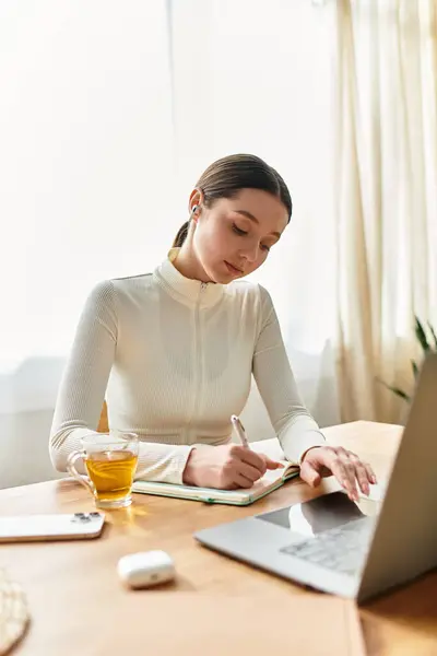 Bella giovane donna si concentra sul suo lavoro da casa, scrivendo note accanto al suo computer portatile. — Foto stock