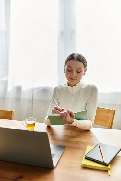 In a cozy kitchen, a young woman writes notes while working on her laptop, sipping tea. — Stock Photo