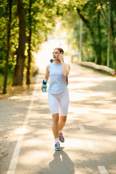 Jogging through a sunny forest path, a young and athletic woman stays hydrated while exercising. — Stock Photo