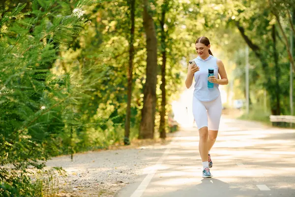 Sporty woman walks in a green park, sipping water and using her phone while exercising. — Stock Photo