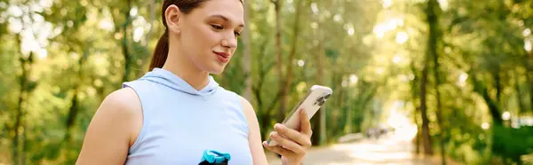 Молодая женщина в спортивной одежде пользуется смартфоном в солнечном парке с деревьями. — стоковое фото