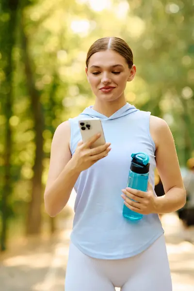 A young athletic woman walks in a sunny park, checking her phone while holding a water bottle. — Stock Photo