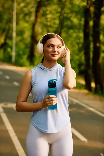 A fit woman walks on a sunny path, enjoying her workout with headphones and water. — Stock Photo