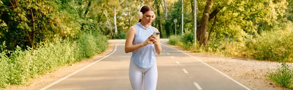 Sunlight shines on a young woman jogging on a peaceful path, checking her phone. — Stock Photo