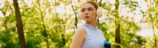 Young athletic woman wears headphones and holds a water bottle, ready for exercise in the park. — Stock Photo