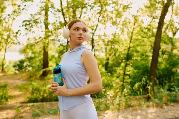 A confident young woman stands gracefully, holding a water bottle and listening to music. — Stock Photo