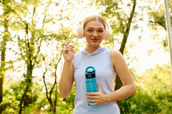 Молода красива жінка посміхається, тримаючи пляшку води, насолоджуючись її тренуванням на природі. — стокове фото