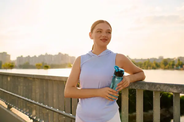 Young athletic woman stands by the water with a smile, holding a water bottle after exercising. — Stock Photo