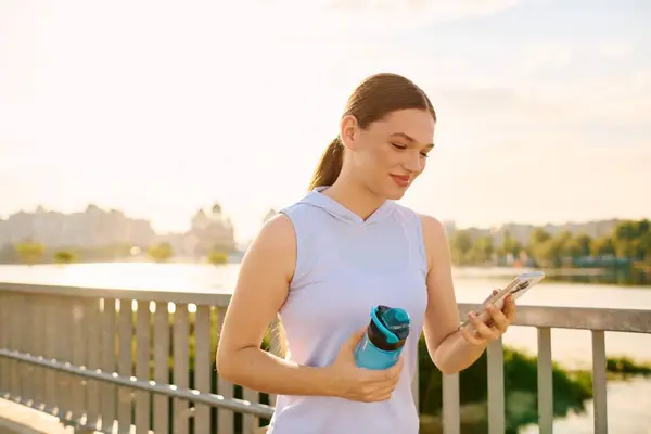 A happy sporty woman by the river holds a water bottle and smiles at her phone. — Stock Photo