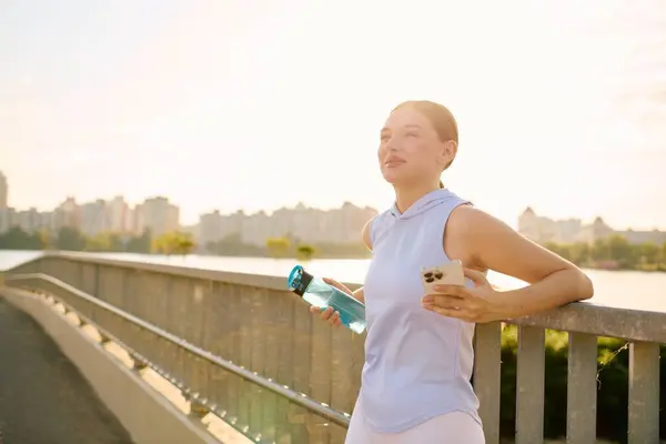 Bella giovane donna si ferma a godersi un drink su un ponte che domina la città al tramonto. — Foto stock